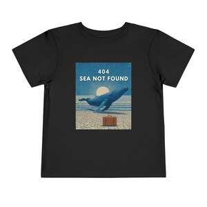 以下が含まれることがあります： 黒い幼児用Tシャツ。海面から飛び出す青いクジラのグラフィックが特徴です。画像にはスーツケースと白い文字で「404 SEA NOT FOUND」と書かれています。デザインは青とベージュの背景に設定されています。