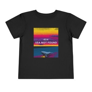 以下が含まれることがあります： 黒い幼児用Tシャツ。クジラのイラストと「404 SEA NOT FOUND」の文字がプリントされています。グラフィックは、黄色、ピンク、紫の色合いの夕焼けのような背景です。クジラはグレーで、海は青色です。