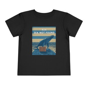 以下が含まれることがあります： 黒い幼児用Tシャツ。青いクジラがスーツケースを持って桟橋に立っているグラフィック。グラフィックには、白文字で「404 SEA NOT FOUND」と青と黄色のストライプの背景が含まれています。