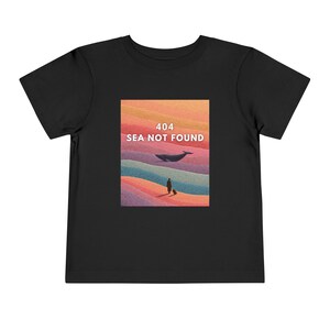 以下が含まれることがあります： クジラ、荷物を持った人、白い文字で「404 SEA NOT FOUND」が描かれた黒い幼児用Tシャツ。グラフィックは、オレンジ、紫、青の色合いを持つカラフルな風景デザインです。