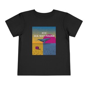Op de afbeelding: Zwart peuter T-shirt met een afbeelding van een walvis, een roze zon en de tekst "404 SEA NOT FOUND". Het ontwerp is geplaatst tegen een achtergrond van een gebarsten aarde en een gradiënthemel.