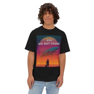 May include: Black t-shirt featuring a graphic design with a whale, a person, and the text "404 SEA NOT FOUND" against a sunset backdrop. The design incorporates shades of orange, purple, and blue.