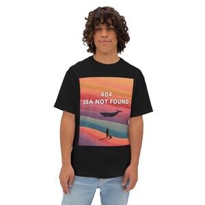 Może przedstawiać: Czarny t-shirt z kolorową grafiką z napisem "404 SEA NOT FOUND". Grafika przedstawia wieloryba, osobę i pofałdowane wzgórza w odcieniach pomarańczu, czerwieni i niebieskiego. Odzież codzienna.