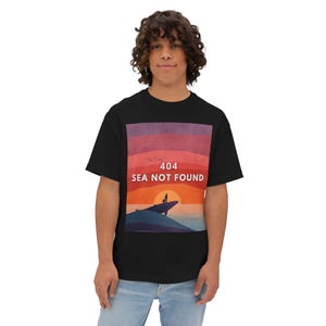 Może przedstawiać: Czarny t-shirt z graficznym wzorem przedstawiającym zachód słońca nad morzem, sylwetkę dwóch osób i biały napis "404 SEA NOT FOUND". Projekt ma odcienie czerwieni, pomarańczy i niebieskiego.