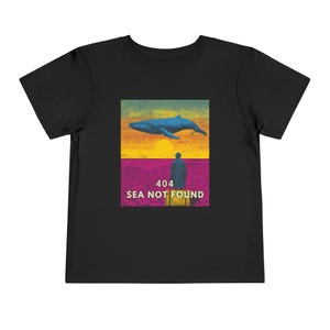 以下が含まれることがあります： グラフィックデザインが施された黒い幼児用Tシャツ。デザインは、夕焼けを背景に、人物の上に青いクジラが描かれています。「404 SEA NOT FOUND」の文字が人物の下にあります。