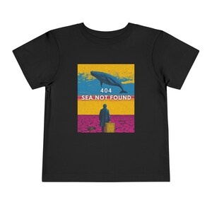 以下が含まれることがあります： 黒い幼児用Tシャツ。クジラ、人物、そして「404 SEA NOT FOUND」のテキストを含むグラフィックデザイン。青、黄、ピンク、黒の色使いでレトロなスタイルです。