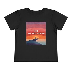 以下が含まれることがあります： 「404 SEA NOT FOUND」のテキストと夕焼けのシーンが描かれた黒い幼児用Tシャツ。 海を見下ろす崖の上に立つ人物と、赤、オレンジ、紫のグラデーションの空がデザインされています。
