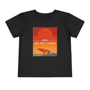 以下が含まれることがあります： 夕焼けの下のビーチにいるクジラを描いたグラフィックが特徴の黒い幼児用Tシャツ。「404 SEA NOT FOUND」の文字が白で表示されています。