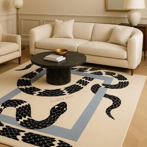 Può includere: Un tappeto beige con un disegno di serpente bianco e nero all'interno di un quadrato azzurro. Il tappeto ha il testo "ZAINAB RUG" al centro. Il tappeto si trova in un soggiorno con un divano e un tavolino.
