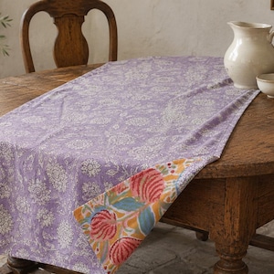 May include: A rectangular table runner featuring a lavender fabric with a white floral pattern on the top side. The corner is folded over to reveal a contrasting underside with a vibrant floral print in shades of pink, orange, and green.