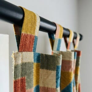 Peut inclure: Un panneau de rideau présentant un motif patchwork composé de blocs de tissu rectangulaires dans des tons jaune moutarde, orange terre cuite, vert sauge et bleu sourd. Le rideau est suspendu à une tringle noire mate par des pattes.