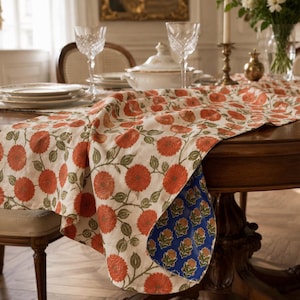 May include: A rectangular table runner featuring a cream-colored base with a repeating pattern of orange flowers and green stems. The underside of the runner is visible, displaying a contrasting blue fabric with a smaller orange floral print pattern.