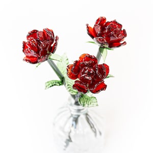 Puede incluir: Un jarrón de cristal transparente contiene tres rosas rojas de cristal con tallos y hojas verdes. Las rosas son de un rojo intenso y brillante, y el jarrón tiene un diseño texturizado y acanalado. El fondo es blanco liso.