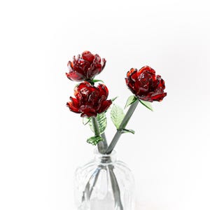 Puede incluir: Un jarrón de cristal transparente contiene tres rosas rojas de cristal con tallos y hojas verdes. El jarrón tiene un diseño texturizado y acanalado. Las rosas son de un color rojo intenso y las hojas son de color verde claro.