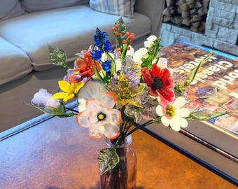 May include: A colorful glass flower bouquet in a pink glass vase. The bouquet features red, white, blue, orange, and yellow flowers with green leaves. The vase sits on a brown table, with a book and a couch in the background.