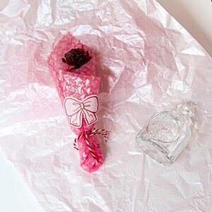 Puede incluir: Una rosa roja oscura envuelta en plástico de burbujas rosa con una pegatina de lazo que dice "love sweet". Una botella de vidrio transparente está al lado de la rosa envuelta sobre un fondo de papel rosa.