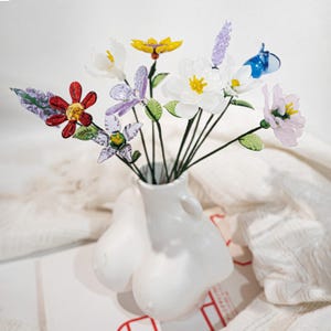 May include: A white ceramic vase in the shape of a torso holds a bouquet of colorful glass flowers. The flowers include red, yellow, purple, and white blossoms with green stems and leaves. The vase has a smooth, glossy finish.