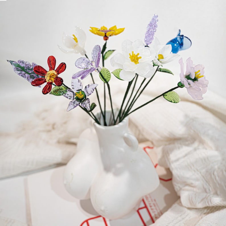 May include: A white ceramic vase in the shape of a torso holds a bouquet of colorful glass flowers. The flowers include red, yellow, purple, and white blossoms with green stems and leaves. The vase has a smooth, glossy finish.