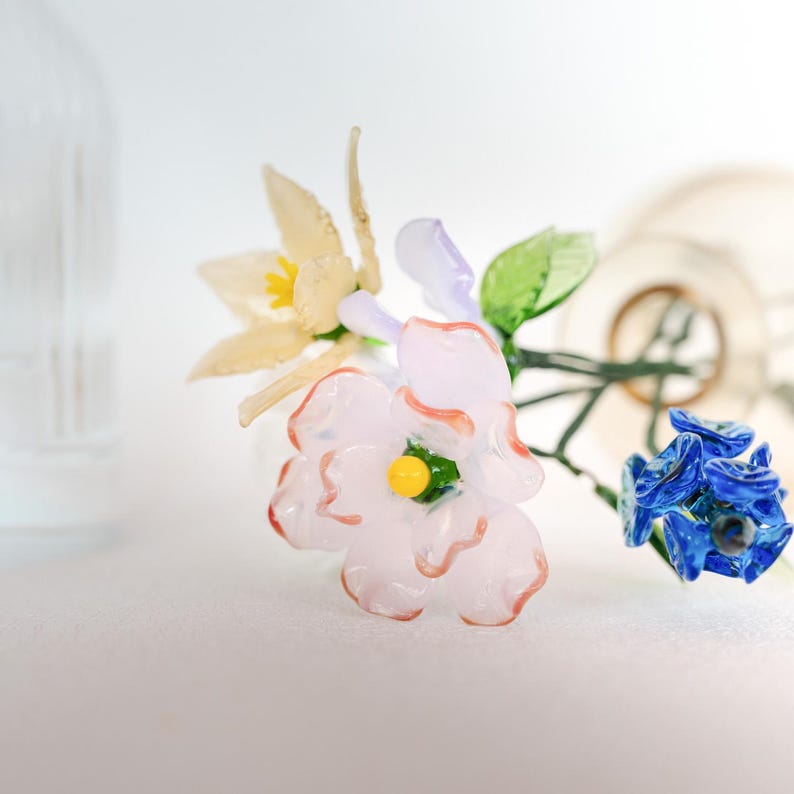 May include: A close-up of a glass flower arrangement featuring a light pink flower with orange accents, a yellow and white flower, and a blue flower. The flowers have green leaves and are set against a white background.
