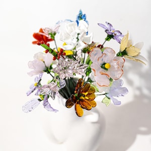 May include: A bouquet of colorful glass flowers in a white vase. The arrangement includes various flower types in shades of red, orange, yellow, purple, and white. The flowers have delicate details and are arranged in a full, eye-catching display.