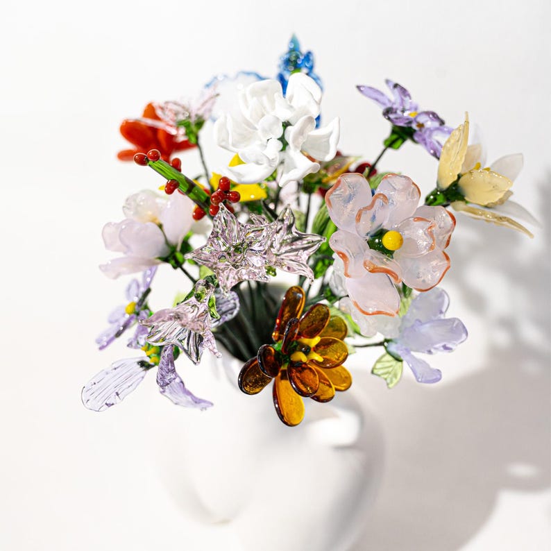 May include: A bouquet of colorful glass flowers in a white vase. The arrangement includes various flower types in shades of red, orange, yellow, purple, and white. The flowers have delicate details and are arranged in a full, eye-catching display.