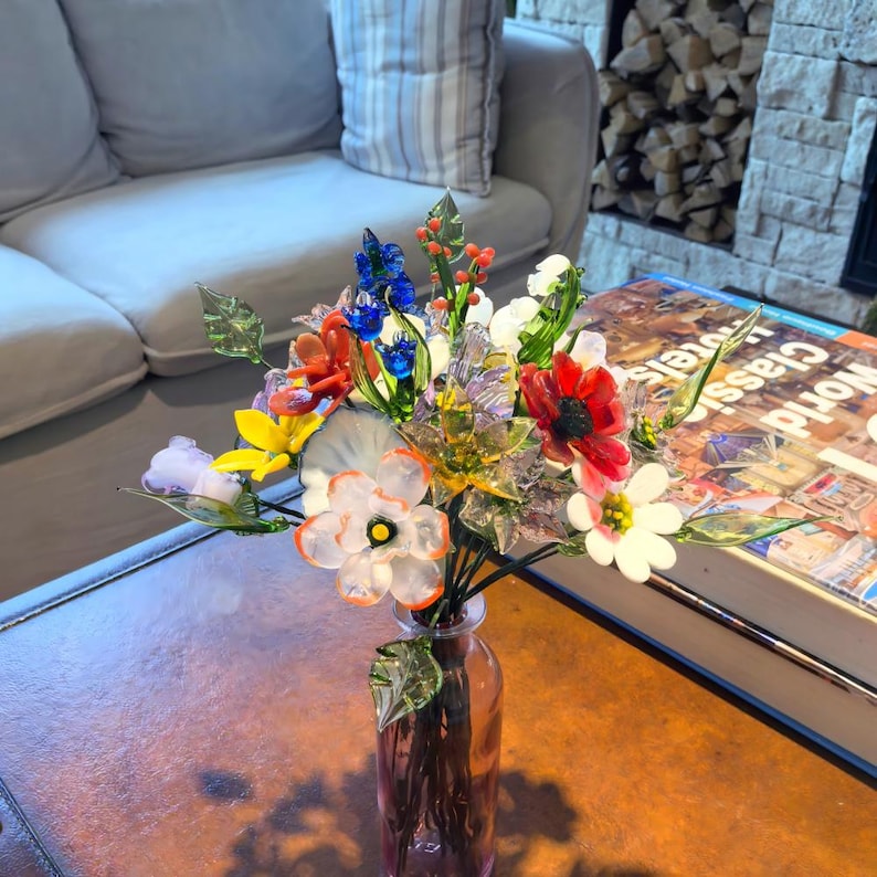 May include: A colorful glass flower bouquet in a pink glass vase. The bouquet features red, white, blue, orange, and yellow flowers with green leaves. The vase sits on a brown table, with a book and a couch in the background.