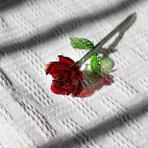 Puede incluir: Una rosa de cristal hecha a mano con una flor de color rojo intenso y hojas verdes. El tallo es de color plateado. La rosa está sobre una superficie blanca y texturizada, con sombras.