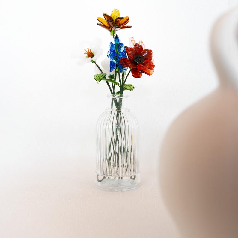 May include: A clear glass vase with vertical ridges holds a colorful bouquet of glass flowers. The flowers include red, blue, white, and yellow blooms with green stems and leaves. The vase sits on a light-colored surface against a white background.