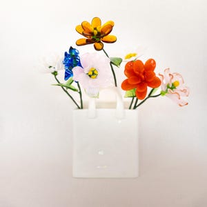 May include: A white ceramic vase shaped like a shopping bag holds a colorful arrangement of glass flowers. The flowers include orange, pink, blue, and white blossoms with green stems. The vase has two handles and sits against a white background.
