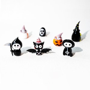 以下が含まれることがあります： ハロウィーンをテーマにしたミニチュアガラスフィギュアのコレクション。鎌を持った黒い死神、赤い帽子をかぶったコウモリ、お化け、カボチャ、魔女の帽子が含まれています。フィギュアは黒、オレンジ、白、ピンクなど、さまざまな色があります。