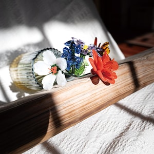 May include: A glass vase with a collection of colorful glass flowers. The flowers include a large white flower with an orange center, blue, red, and purple flowers. The vase is resting on a wooden surface.