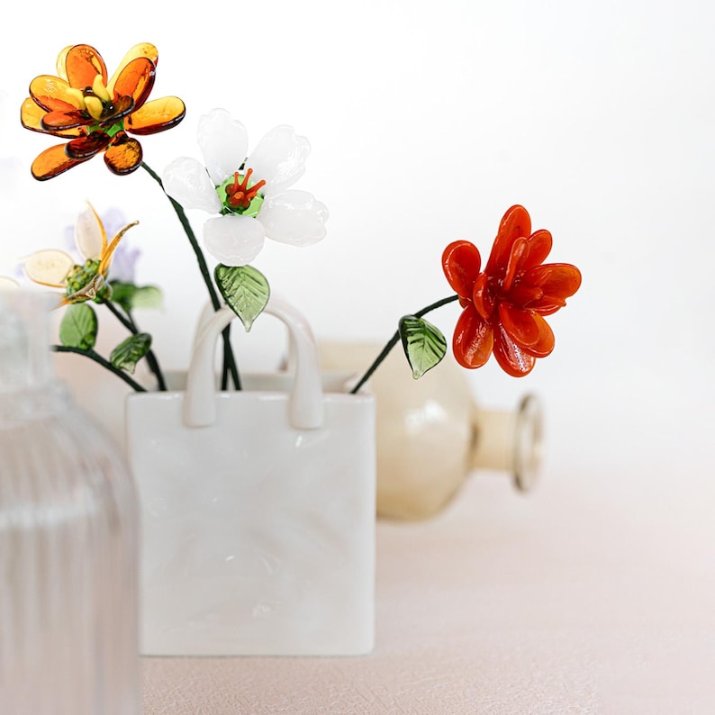 May include: A white ceramic shopping bag vase holds colorful glass flowers. The flowers include orange, white, and red blooms with green leaves. The vase is on a light pink surface.
