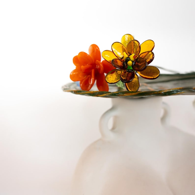 May include: Two glass flowers, one orange and one yellow-brown, rest on a decorative glass plate. The flowers are detailed with multiple petals and sit atop a white vase with two handles. The background is a soft white.