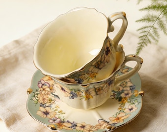 May include: Two cream-coloured teacups and a saucer with floral designs. The teacups have gold trim and ornate handles. The saucer is square with a scalloped edge and a pale blue border. The floral pattern includes purple, yellow, and blue flowers.