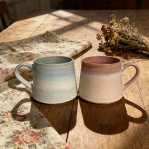 May include: Two ceramic mugs with curved handles. One mug is light blue and white, the other is pink and white. The mugs are on a wooden table with a floral patterned cloth and dried flowers.