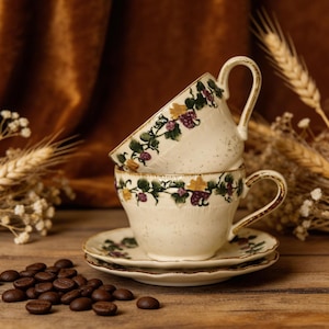 Könnte beinhalten: Zwei cremefarbene Teetassen mit passenden Untertassen, verziert mit einem Weinreben-Design in Grün, Lila und Gelb. Kaffeebohnen sind auf einer Holzoberfläche verstreut, mit einem braunen Samthintergrund.