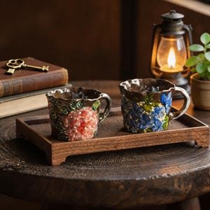 May include: Two decorative ceramic mugs with floral designs, one with red flowers and the other with blue flowers, sit on a wooden tray. The mugs have brown rims and handles. A vintage key and book are in the background.