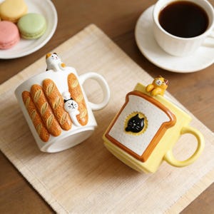 Puede incluir: Dos tazas de cerámica con diseños de gatos y comida. Una taza es blanca con detalles de baguette y una figura de gato. La otra es amarilla con un diseño de tostada y una figura de gato. Un plato de macarons y una taza de café están en el fondo.