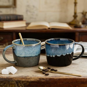 Könnte beinhalten: Zwei Keramikbecher mit blau-schwarzer Ombre-Glasur. Ein Becher hat einen goldenen Löffel im Inneren. Die Becher stehen auf einer Holzoberfläche mit Zuckerwürfeln, Kaffeebohnen und einem goldenen Löffel. Die Becher haben einen Henkel.
