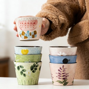 May include: A collection of hand-painted ceramic bowls with floral patterns. The bowls display a range of colours, including pink, blue, green, and white, with illustrations of flowers and leaves. The bowls are stacked, with one bowl being held up.