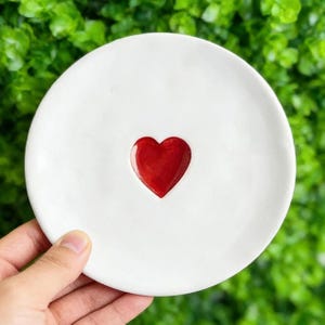 May include: A small, white ceramic dish with a glossy, red heart-shaped indentation in the center. The dish is round and appears handmade. The background is blurred green foliage.