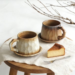 May include: Two ceramic mugs with matching saucers. One mug is off-white with brown speckles, the other is brown. A slice of cake and a fork sit on a white cloth. The mugs have a textured, faceted design.