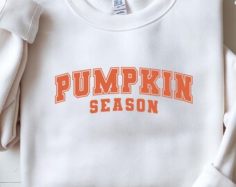 May include: A white sweatshirt with the words "PUMPKIN SEASON" printed in orange block letters. The sweatshirt has a crew neck and long sleeves. The text is in a collegiate style font.