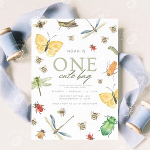 Pode incluir: Um cart&atilde;o de convite branco com ilustra&ccedil;&otilde;es em aquarela de insetos, incluindo borboletas, abelhas e lib&eacute;lulas. O texto diz "NOAH IS ONE cute bug" com detalhes da festa. Fita azul clara e uma bobina de madeira no fundo.