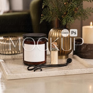 May include: A candle jar with a blank label, a small glass bottle of matches, and a black candle snuffer sit on a marble tray. A gold vase with greenery and a lit candle are in the background.
