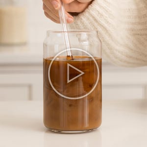 May include: A clear glass can-shaped tumbler filled with a brown liquid, likely coffee or a similar beverage. A clear straw is inserted into the drink. The drink is being stirred. The glass sits on a white surface.