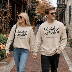 May include: Two people wearing matching beige sweatshirts with the text "Ladybug Mocks" in black script. The sweatshirts are paired with blue jeans and black trousers. They are walking down a brick-lined street.