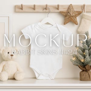 May include: A white baby onesie hangs on a white hanger. The word "MOCKUP" is displayed above the text "RABBIT SKINS 4424". A teddy bear, a wooden star, a small Christmas tree, and a stocking are also in the image.
