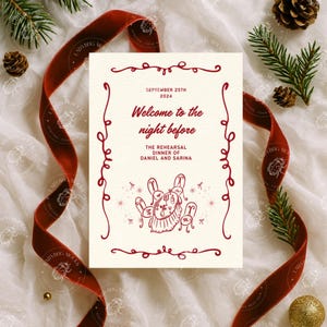 May include: A cream-colored invitation card with red text and illustrations, featuring the words "Welcome to the night before" and details for a rehearsal dinner. The card is surrounded by a red velvet ribbon, pine needles, and decorative elements.
