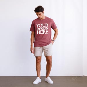 May include: A maroon t-shirt with the text "YOUR DESIGN HERE" in white block letters. The shirt is paired with khaki shorts and white sneakers. The person is wearing a watch.